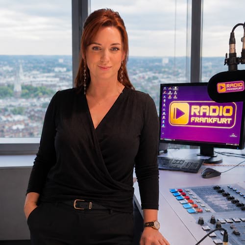Frau in schwarzer Bluse steht vor einem Radiostudio mit einem Computer und Mischpult, Blick aus dem Fenster auf die Stadt.