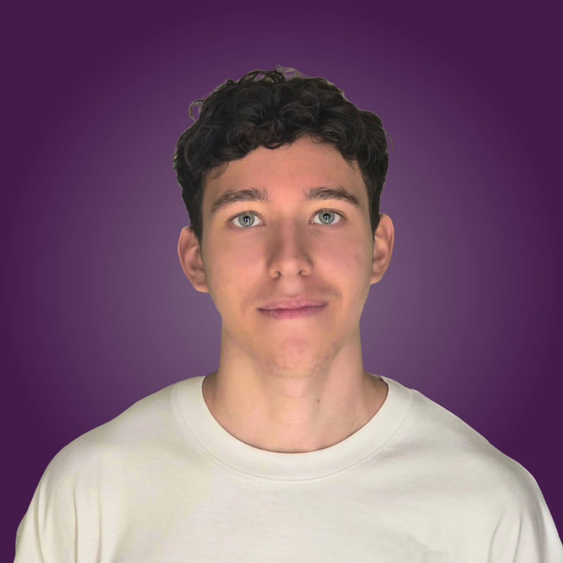 Junger Mann mit lockigem, dunklem Haar und blauen Augen, trägt ein helles T-Shirt vor lila Hintergrund.