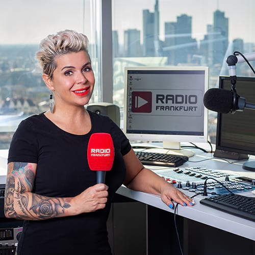 Frau mit kurz geschnittenen, blonden Haaren und tätowierten Armen hält ein Mikrofon in einem Radiostudio mit Stadtansicht.