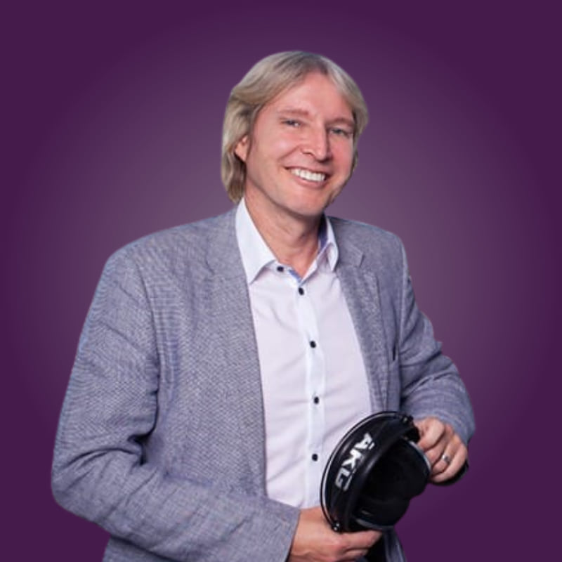 Lächelnder Mann mit blonden Haaren in einem grauen Blazer hält Kopfhörer in der Hand. Hintergrund lila.