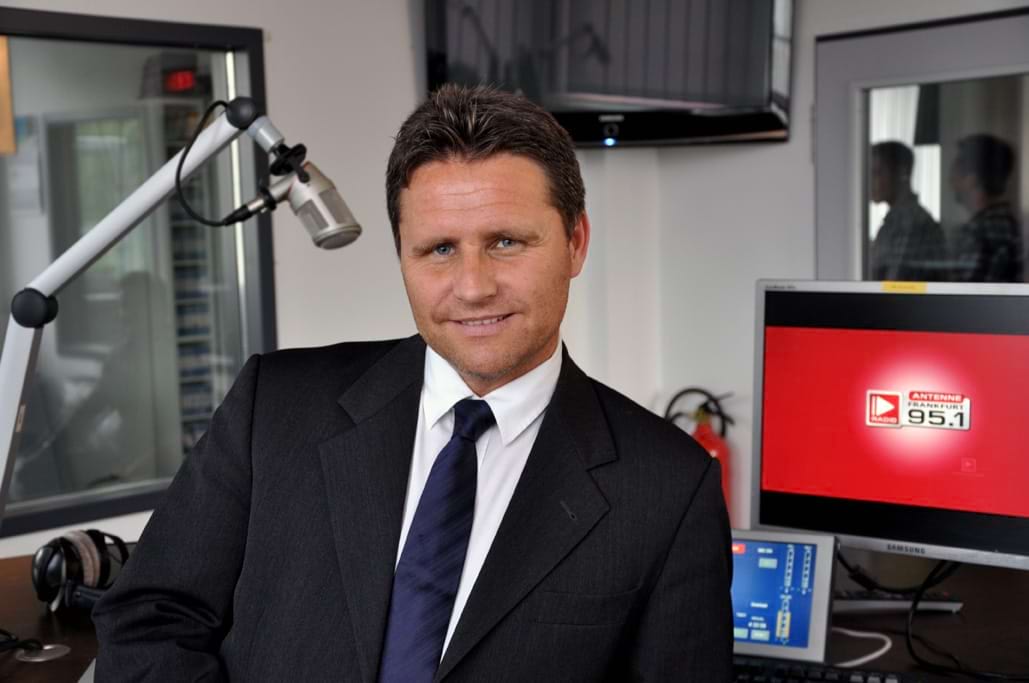 Mann im Anzug mit lässigem Lächeln in einem Studio, Mikrofon und Bildschirm im Hintergrund sichtbar.